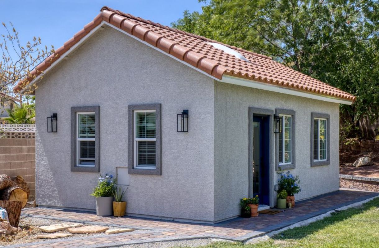 White casita in backyard with red roof, a few plants set outside